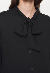 Femme portant un chemisier noir à boutons avec un grand nœud noué au niveau du col, laissant apparaître son cou et une partie de ses cheveux.