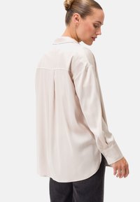 Blusa beige chiaro dal fit rilassato, maniche lunghe e orlo curvo. Presenta una piega sul retro e polsini con bottone per un tocco in più.