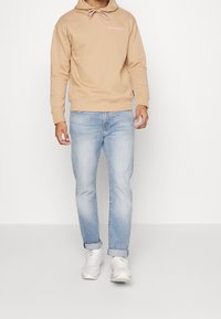 Felpa con cappuccio color cammello con logo rosa, abbinata a jeans azzurri e sneakers bianche. Tessuto liscio, taglio casual, risvolti sui jeans.