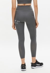 Graue hoch taillierte Leggings mit glatter Textur, die perforierte Akzente an den Unterschenkeln und ein Logo-Detail auf der Rückseite aufweisen.