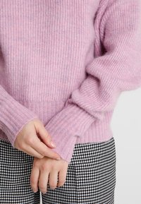 Suéter de color rosa claro de canalé en material suave y esponjoso, con mangas largas y un corte holgado. Combinado con pantalones de pata de gallo en blanco y negro.