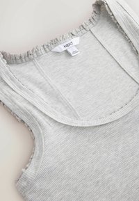 Light gray ribbed tank top with delicate ruffled edges around the neckline and armholes, featuring a white brand label reading "NEXT".
