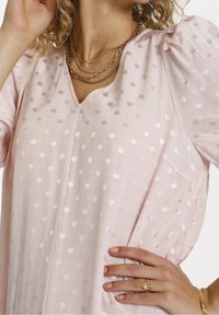 Světle růžová blůza s jemnými bílými puntíky, s nafouknutými rukávy a výstřihem do V; má přední kapsu a hladkou, strukturovanou tkaninu.