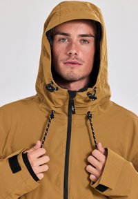 Bruine waterdichte jas met een capuchon, zwarte voering, trekkoordverstellingen en klittenbandsluitingen. Voorzien van een rits aan de voorkant en subtiele branding.