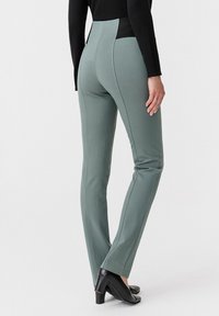Pantaloni aderenti a vita alta in verde tenue, realizzati in tessuto liscio e con cuciture verticali, abbinati a tacchi neri e a un top nero aderente.
