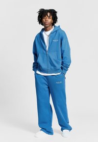 Modrá mikina s kapucí na zip a sladěné tepláky vyrobené z měkkého materiálu, s uvolněným střihem a vyšitými logy. Bílé tričko pod tím a boty.