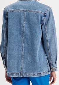 Veste en denim bleu clair avec une coupe droite, des poignets à boutons et un col classique. Présente des coutures visibles et une texture lisse.