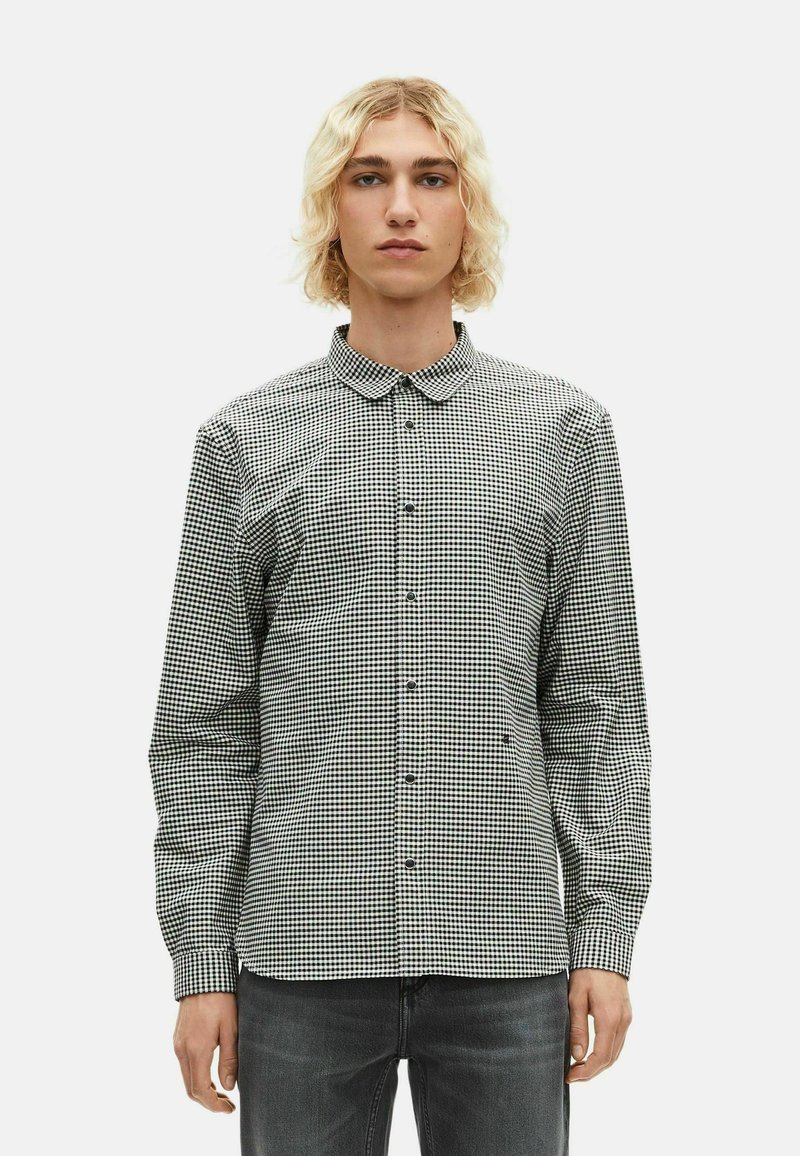 Pessoa jovem com cabelo loiro até aos ombros, vestindo uma camisa de botões em padrão xadrez preto e branco e jeans cinza escuro, de pé contra um fundo simples.