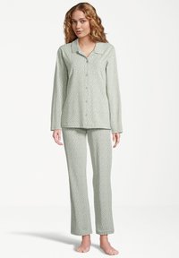 Nainen seisoo paljain jaloin, puettuna vaaleanvihreään pitkähihaiseen napilliseen pyjaman yläosaan ja siihen sopiviin housuihin, joissa on pieniä valkoisia pilkkuja valkoisella taustalla.