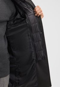 Cappotto nero con fodera interna grigia, caratterizzato da un motivo trapuntato. Include tasche e una chiusura con zip sul lato destro.