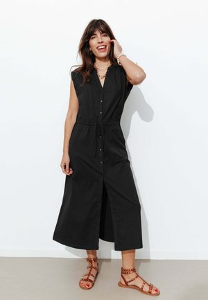 Femme souriante debout, portant une robe midi noire sans manches à boutons avec une ceinture à la taille et des sandales marron cloutées.