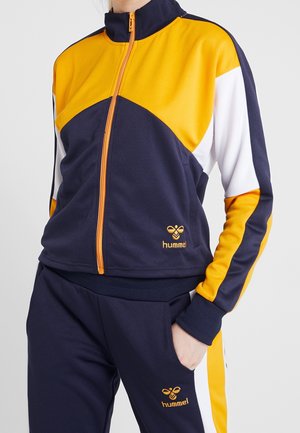 Kvinde iført en marineblå, gul og hvid lynlåssportjakke og matchende bukser med hummel-logo broderet på begge.