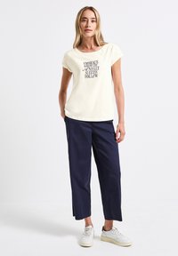 T-shirt en coton crème avec texte graphique noir, associée à un pantalon ample bleu marine et des baskets blanches. Design simple et épuré avec un style décontracté.