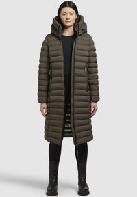 Polstret lang frakke i olivengrøn med hætte, featuring en lynlås foran, sidelommer og en glat tekstur. Piping-detaljer langs sømmene.