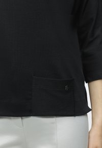 Άτομο που φορά μαύρη μπλούζα με μικρή μπροστινή τσέπη και ανοιχτό γκρι παντελόνι, εμφανίζεται μόνο το κάτω μέρος του κορμού και μέρος του ενός βραχίονα.