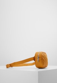 Sac rectangulaire en cuir jaune avec une texture matelassée, fermeture éclair et bandoulière réglable, posé sur un socle blanc sur un fond clair.