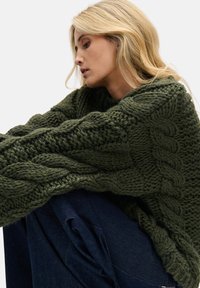 Zielony sweter na drutach z grubymi warkoczami, okrągłym dekoltem i oversize'owymi rękawami, zestawiony z ciemnymi jeansami. Miękki, teksturowany materiał.