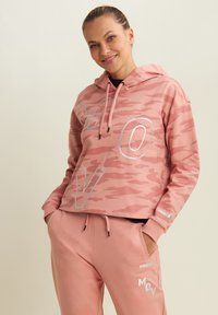 Donna sorridente con un felpa a motivo camouflage rosa con il testo "MOV" e pantaloni della tuta rosa abbinati con il nome del marchio "FREDDY" e il testo "MOV".