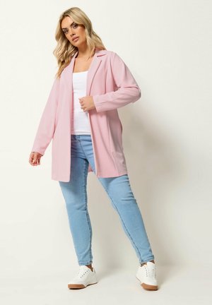 Vaaleatukkainen nainen, jolla on yllään pitkä vaaleanpunainen bleiseri, valkoinen paita, vaaleansiniset farkut ja valkoiset lenkkarit, seisoo yksinkertaisen taustan edessä.