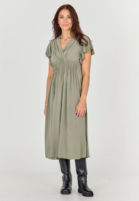 Robe midi vert olive avec manches courtes volantées et taille froncée. Tissu doux et fluide, fentes latérales, associée à des bottes noires hauteur genou.