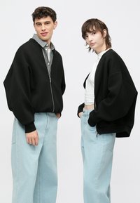 Zwarte oversized bomberjack met ritssluiting, gecombineerd met lichtblauwe jeans met wijde pijpen. Het jack heeft geribbelde manchetten en een losse pasvorm.