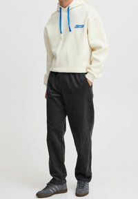 Světle krémová mikina s modrými tkaničkami a logem, spárovaná s tmavě šedými kordovými kalhotami. Šedé tenisky dokreslují celý outfit.