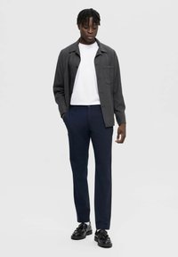 Γκρίζο πουκάμισο με κουμπιά πάνω από λευκό μπλουζάκι, συνδυασμένο με ναυτικές παντελόνες και μαύρα παπούτσια. Το πουκάμισο διαθέτει τσέπη στο στήθος και υφή υφάσματος.