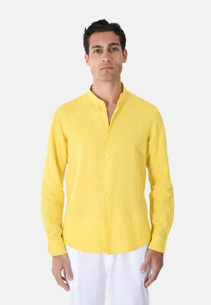 Uomo che indossa una camicia gialla a maniche lunghe con bottoni e colletto alla coreana e pantaloni bianchi, in piedi davanti a uno sfondo semplice.