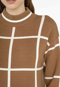 Pull en maille marron avec un col montant, motif à carreaux blanc et tissu texturé. Coupe décontractée avec des épaules tombantes.