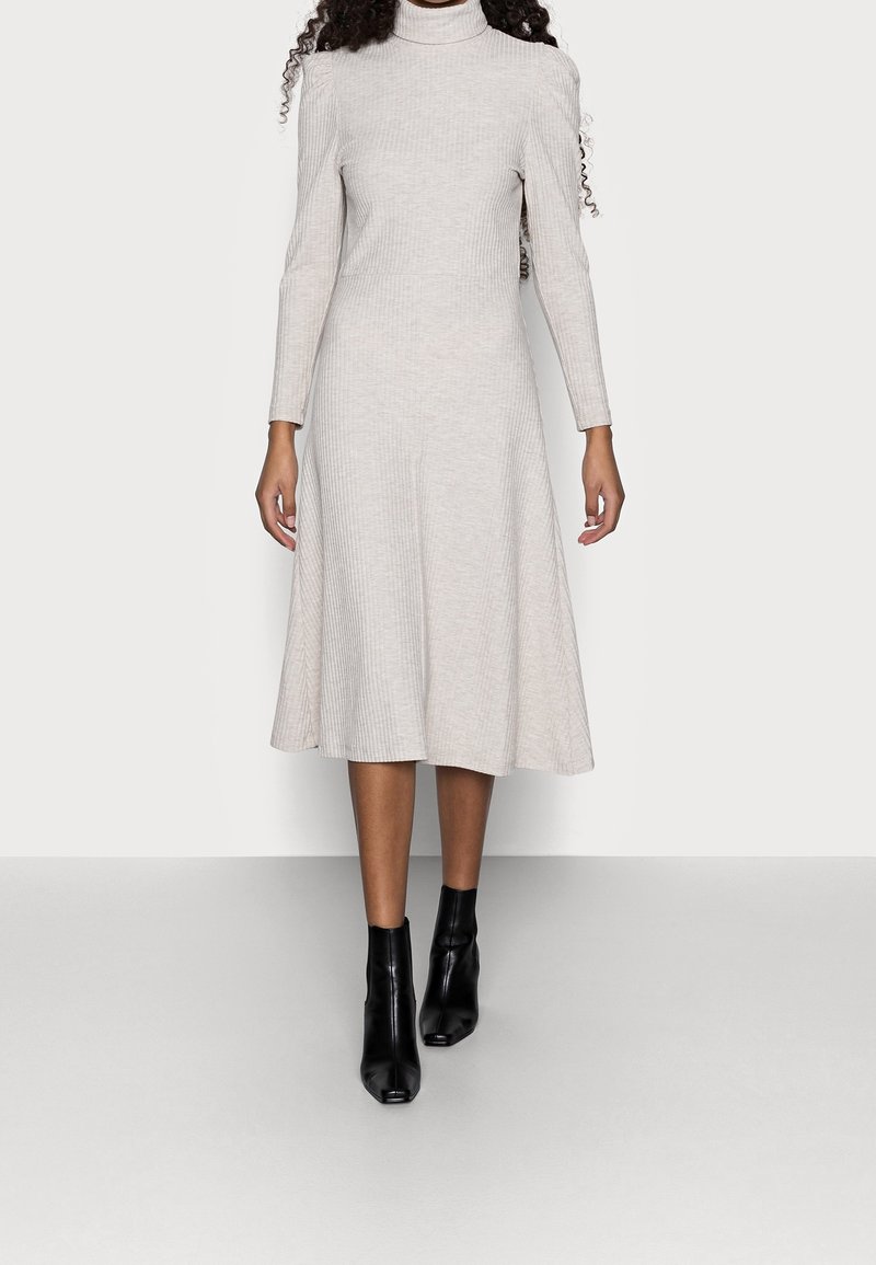 Femme portant une robe midi côtelée beige clair à manches longues avec un col roulé et des bottines noires, debout devant un fond uni.