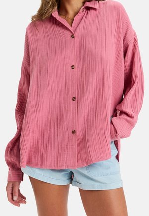 Femme portant une chemise ample rose texturée à boutons avec manches longues et un short bleu clair à ourlet remonté.
