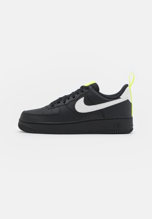Černý teniska Nike Air Force 1 s bílým logem Swoosh a neonově žlutým poutkem a detailem na jazyku, zobrazená z boku na bílém pozadí.