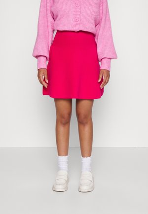 Osoba ubrana w różowy sweter z dzianiny, jasnoróżową spódnicę, białe skarpetki i białe wsuwane buty, stojąca na jednolitym, jasnym tle.