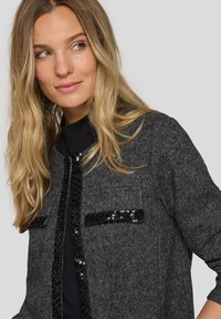 Graue Strickjacke mit schwarzen Paillettenakzenten an Taschen und Vorderöffnung, mit rundem Ausschnitt und strukturierter Stoffqualität.