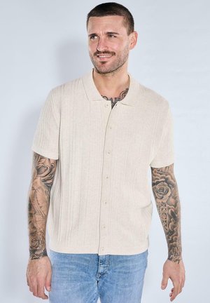 Mann mit kurzen Haaren und Tätowierungen an beiden Armen, trägt ein beigefarbenes kurzärmeliges Hemd mit Knöpfen und blaue Jeans, leicht lächelnd.