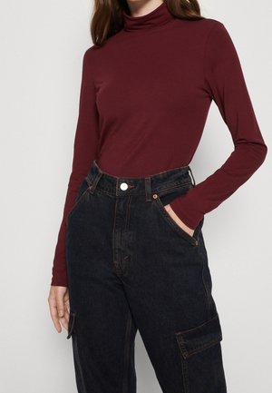 Femme portant un col roulé bordeaux à manches longues rentré dans un jean cargo noir taille haute, une main dans la poche.