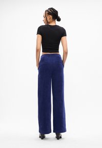 Γυναίκα με πλεξούδες που φοράει ένα στενό μαύρο crop top, ψηλόμεσα παντελόνια από μπλε βελούδο με φαρδιά πόδια και μαύρες γόβες, στέκεται με τα χέρια στα τσέπες.