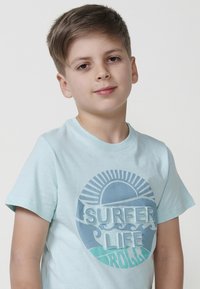 Šviesiai mėlynas medvilninis marškinėlis su apvaliu grafiniu atvaizdu, kuriame užrašas "SURFER LIFE" baltos ir mėlynos spalvose, vaizduojantis bangas ir saulės motyvą.