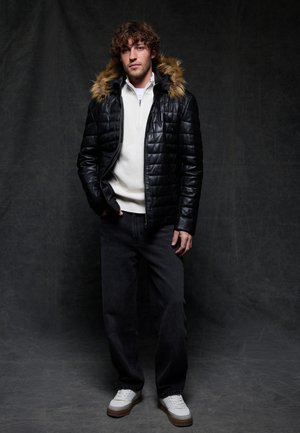 Young man with curly hair wearing a black quilted jacket with fur hood, white quarter-zip sweater, black jeans, and white sneakers against dark background.