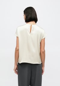 Ανοιχτό μπεζ μπλούζα με κοντά μανίκια και άνοιγμα στην πλάτη. Λείο, λαμπερό ύφασμα με χαλαρή εφαρμογή. Συνδυασμένη με γκρι παντελόνι ψηλής μέσης.