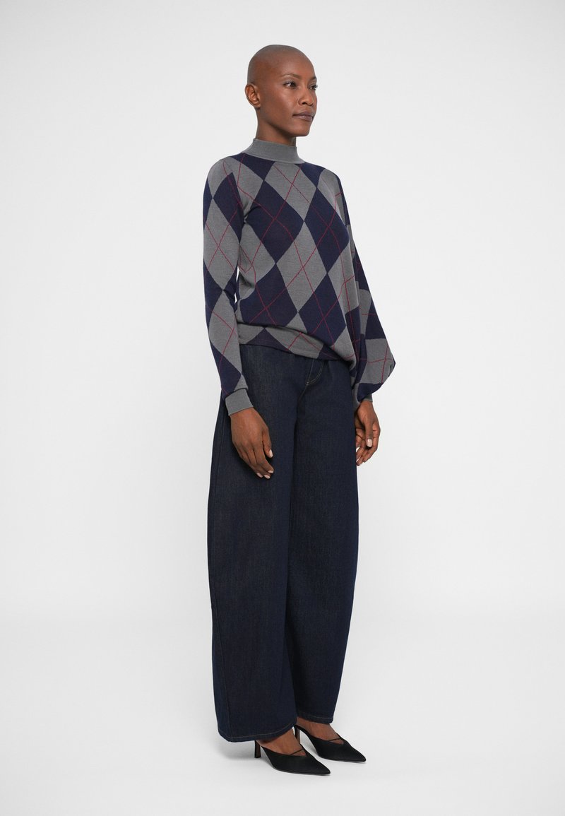 Maglione argyle grigio e navy con colletto alto, abbinato a jeans in denim scuro a gamba larga e décolleté nere a punta. Design semplice e pulito.