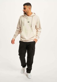 Lys beige hettegenser med "DEF" logo, frontlomme og snorhette, kombinert med svarte cargo-bukser og hvite sneakers.