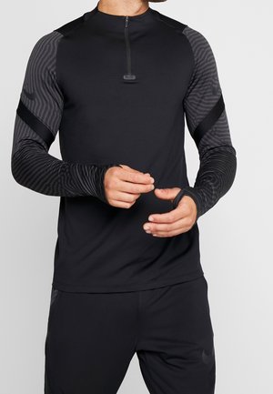 Mężczyzna w czarnym sportowym topie z szarymi, wzorzystymi rękawami z otworami na kciuki, poprawiający rękaw, pokazany od szyi do połowy uda.