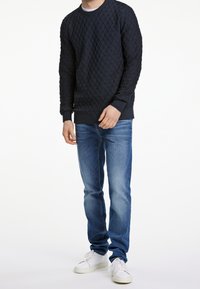 Marineblauer, gesteppter Pullover mit strukturiertem Rautenmuster, kombiniert mit hellblauen Jeans und weißen Sneakers, die einen lässigen Lagenlook präsentieren.