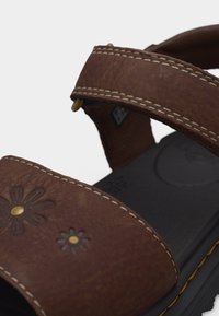 Καφέ δερμάτινο σανδάλι με λουλουδένια κοψίματα και ορείχαλκινα καρφιά, με διπλές λουρίδες και άσπρες ραφές σε μαύρη σόλα με κίτρινες λεπτομέρειες.