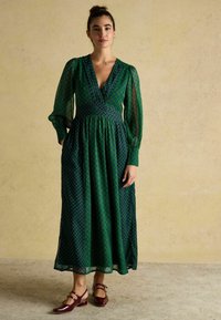 Długa zielona sukienka z wzorzystą teksturą, głębokim dekoltem w kształcie litery V i bufiastymi rękawami. Połączona z burgundowymi butami. Gładki, lejący się materiał.