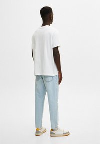 Hombre de espaldas, vistiendo una camiseta blanca sencilla, jeans azul claro y zapatillas blancas con detalles naranjas, frente a un fondo liso.