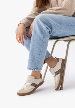 Persona sentada en una silla que lleva pantalones vaqueros azul claro y zapatillas beige con acentos de estampado animal marrón y cordones blancos.