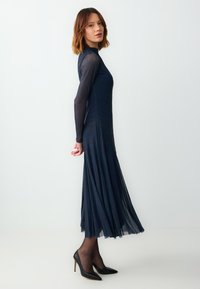 Abito lungo a maniche con colore blu navy, con un corpetto aderente e una gonna fluida a pieghe, realizzato in tessuto trasparente. Abbinato a tacchi neri a punta.