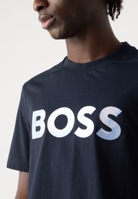 Tamsiai mėlynas medvilninis marškinėliai su dideliu, gradientiniu „BOSS“ logotipu baltos ir šviesiai mėlynos spalvų. Trumpomis rankovėmis, apvaliu iškirpimu, minkšta tekstūra.